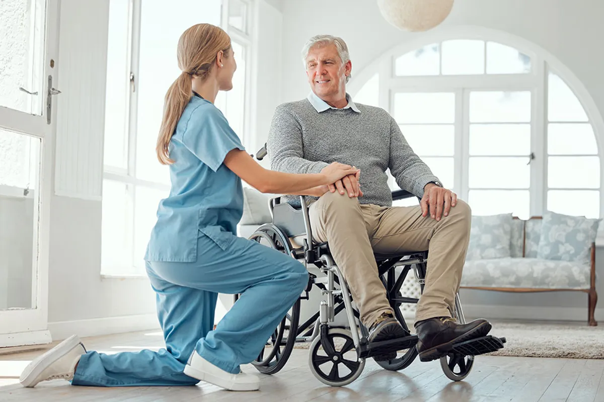 Caregiver kneeling beside an elderly man in a wheelchair at home