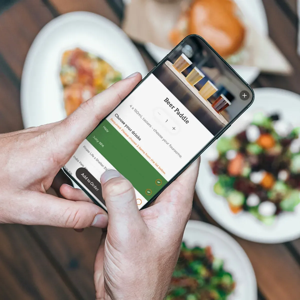 Phone showing a HungryHungry beer paddle order screen above plates of shared food